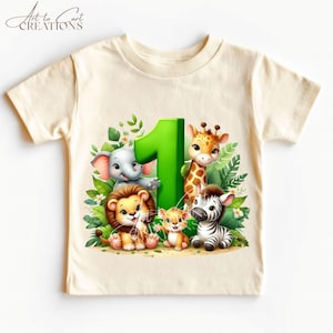 以下が含まれることがあります： ジャングルをテーマにした、クリーム色の幼児用Tシャツ。デザインには、緑色の大きな数字1と、象、ライオン、キリン、シマウマ、2匹の子どもたちの漫画の動物が、緑の葉の背景に囲まれています。