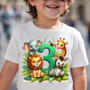 以下が含まれることがあります： ライオン、ゾウ、キリン、シマウマと数字の3が描かれた、白いTシャツ。デザインには緑の葉と「Art to Cart Creations」の文字が含まれています。