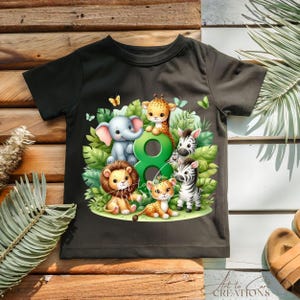 以下が含まれることがあります： 黒いTシャツ。象、キリン、ライオン、シマウマ、ヒョウなど、漫画の動物のカラフルなイラストが描かれています。数字の8と緑の葉と蝶が動物たちを囲んでいます。「Art to Care Creations」の文字が下にあります。