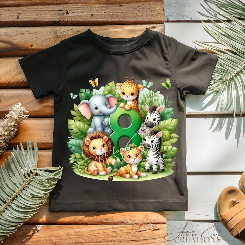 以下が含まれることがあります： 黒いTシャツ。象、キリン、ライオン、シマウマ、ヒョウなど、漫画の動物のカラフルなイラストが描かれています。数字の8と緑の葉と蝶が動物たちを囲んでいます。「Art to Care Creations」の文字が下にあります。