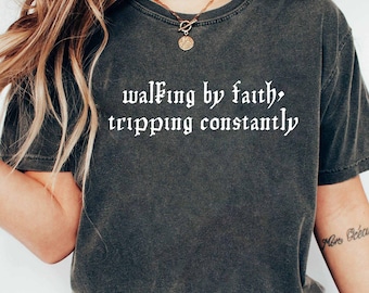 May include: Dark gray t-shirt with the text "walking by faith, tripping constantly" in a white gothic font. The shirt is a crew neck style and appears to be made of a soft, comfortable material. The model is wearing blue jeans.
