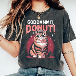 May include: Dark gray t-shirt with the text "GODDAMMIT, DONUT!" above a cartoon cat wearing sunglasses and a crown. The cat is orange and brown, set against a red circle with star accents. A fun, casual graphic tee.