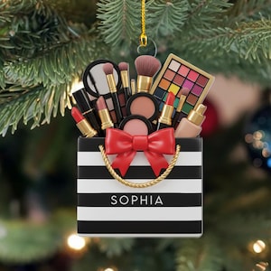 May include: A Christmas tree ornament shaped like a black and white striped shopping bag filled with makeup products. The bag has a red bow and gold rope handles. The name "SOPHIA" is printed on the bag.
