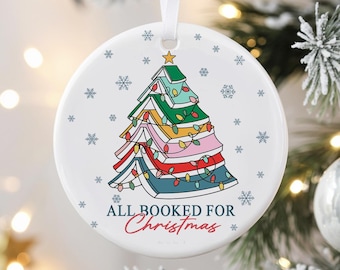 May include: White ceramic Christmas ornament with a colorful book-shaped Christmas tree design. The tree is made of stacked books in various colors, adorned with string lights and a star. Text reads "ALL BOOKED FOR Christmas". Snowflakes surround the design.