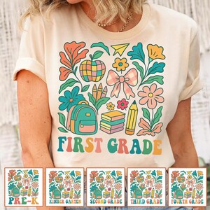 Könnte beinhalten: Ein cremefarbenes T-Shirt mit einem bunten Grafikdruck von Schulmaterialien, darunter ein Rucksack, Bücher, ein Bleistift, ein Apfel, eine Schleife und Blumen. Der Text "FIRST GRADE" ist in großen, mehrfarbigen Retro-Buchstaben aufgedruckt.