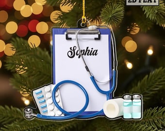 May include: A medical-themed Christmas ornament featuring a blue clipboard with the name "Sophia", a blue stethoscope, pills, bandages, and vials. The ornament is hanging in front of a Christmas tree with bokeh lights.