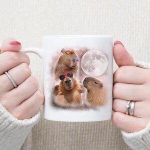 May include: White ceramic mug with a colorful illustration of three capybaras, one wearing sunglasses, another with a leaf on its head, and a full moon in the background.