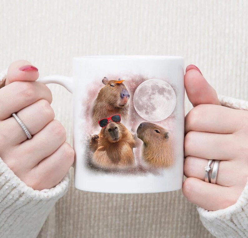 May include: White ceramic mug with a colorful illustration of three capybaras, one wearing sunglasses, another with a leaf on its head, and a full moon in the background.
