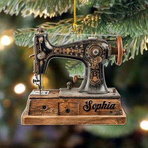 May include: A black and gold sewing machine ornament with the name "Sophia" on a wooden base. The ornament features detailed floral patterns and a wooden wheel. It is suspended by a gold string, suitable for festive decoration.