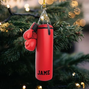 May include: A red boxing-themed ornament featuring a miniature punching bag and boxing gloves. The punching bag has the name "JAME" printed on it. The gloves are red and black. The ornament hangs from a silver chain.