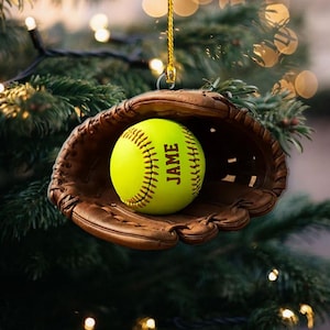 May include: A softball Christmas ornament featuring a neon yellow softball with the name "JAME" printed on it, nestled inside a brown leather baseball glove. The ornament hangs from a gold string.