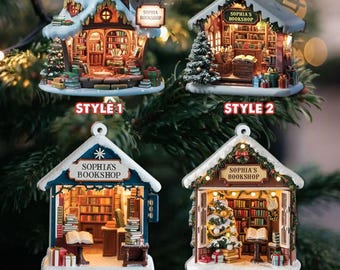 May include: Four different styles of Christmas ornaments shaped like a small bookstore with a sign that reads "Sophia's Bookshop". The ornaments are decorated with lights, snow, and a small Christmas tree.