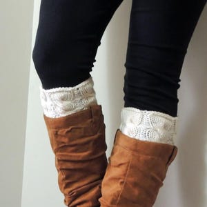 May include: A pair of off-white, cable-knit boot cuffs featuring a textured, circular pattern. The cuffs are worn over black leggings and tucked into brown, slouchy, mid-calf suede boots, creating a layered look with the knit fabric.