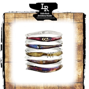 Op de afbeelding: Een set van vijf zilveren golfarmbanden met verschillende gekleurde metalen inlays. De armbanden zijn op elkaar gestapeld en zijn gerangschikt in een golfpatroon. De armbanden zijn gemaakt door Little Rock Jewellery Studio.