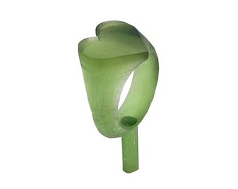 May include: A translucent light green ring featuring a smooth, rounded band and a flat, heart-shaped top surface. The ring is positioned vertically, supported by a small, matching green cylindrical base extending from the bottom.