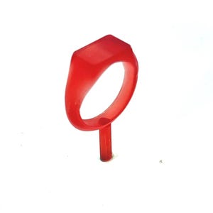May include: A vibrant red ring with a rectangular top, set on a thin, matching stem. The ring's design is simple and modern, with a smooth, polished surface. The ring is likely a piece of jewelry or a fashion accessory.
