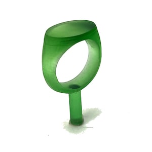 May include: A translucent green ring with an oval face and a rounded band. The ring is attached to a cylindrical stem, likely for display or manufacturing purposes. The ring's design is simple and modern.