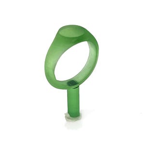 Op de afbeelding: Een doorschijnende groene ring met een afgeronde bovenkant, gemonteerd op een groene paal. De ring heeft een effen kleur en lijkt van een glad materiaal te zijn gemaakt. De ring is waarschijnlijk een sieraad.