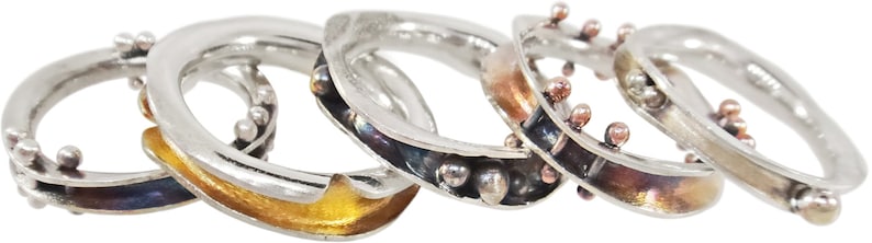 May include: Five silver rings with different coloured accents. The rings are stacked on top of each other. The accents include gold, copper, and black.