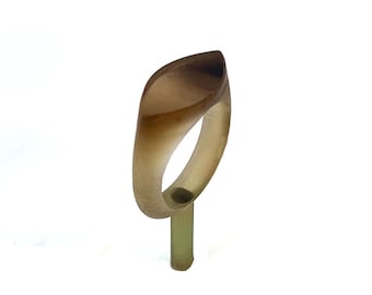 May include: A unique, abstract ring with a leaf-like design. The ring features a translucent, light brown hue with darker brown accents. The ring is mounted on a light green stand, showcasing its form against a white background.