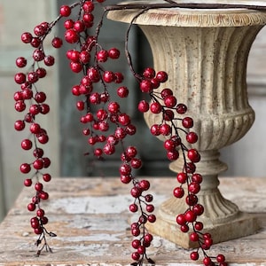 May include: Red berry stems with a glittery finish cascade down, alongside a decorative urn. The berries are a deep red color, and the stems are dark brown. The urn is a light beige color, with a textured surface. The arrangement is set on a distressed white wooden surface.