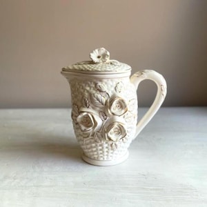 May include: Cream-colored ceramic pitcher with a lid, featuring embossed roses and leaves. The pitcher has a handle and a basketweave texture. The roses are accented with gold trim, adding a touch of elegance to this decorative piece.
