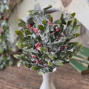 May include: A festive arrangement of faux mistletoe in a white vase. The green leaves are dusted with white, and bright red berries add a pop of color. This decorative piece is perfect for holiday decor.