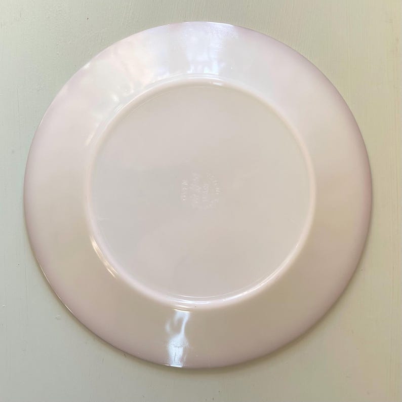 May include: The underside of a white ceramic plate with a pink rim. The plate has a small, circular mark in the centre that reads "Fire King USA".