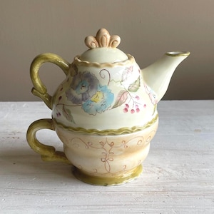 May include: A cream-colored ceramic teapot and teacup set. The teapot features a floral design with blue, purple, and pink flowers, and a decorative finial on the lid. The teacup is integrated into the base of the teapot, with a matching floral pattern and a green handle.