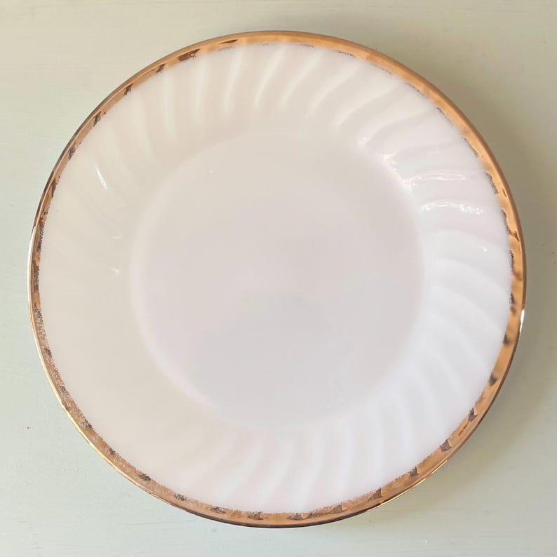 May include: A white milk glass dinner plate with a gold rim. The plate has a swirl design.