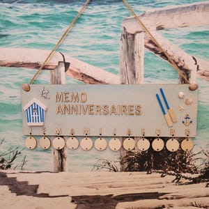 Peut inclure: Un tableau mémo en bois bleu clair avec les mots "MEMO ANNIVERSAIRES" et les mois de l'année. Le tableau est décoré d'éléments nautiques, dont une petite maison, des rames et une ancre. Le tableau est suspendu par une corde.