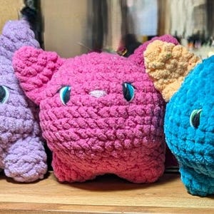 May include: Three crocheted plush toys shaped like cats. The cat on the left is purple, the middle cat is pink, and the cat on the right is blue with orange accents.