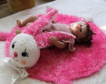 May include: A pink, fluffy bunny-shaped baby mat with a white, crocheted bunny head and a satin bow. A baby doll is lying on the mat, wearing a pink outfit. The mat is on a white, fluffy rug.