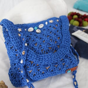 May include: A blue crochet shoulder bag with a flap closure, adorned with seashells and star-shaped embellishments. The bag features a woven pattern and a long strap, perfect for carrying essentials. The bag is approximately 10 inches wide.