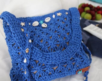 May include: A blue crochet shoulder bag with a flap closure, adorned with seashells and star-shaped embellishments. The bag features a woven pattern and a long strap, perfect for carrying essentials. The bag is approximately 10 inches wide.