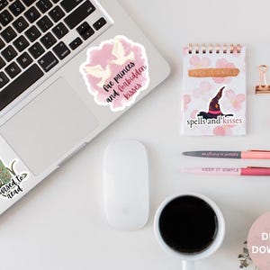 May include: A flat lay of a laptop with stickers on it. The stickers are pink and white and have text that says "buy me books", "cursed to read", "fae princes and forbidden kisses", and "spells and kisses". There is also a white mouse, a cup of coffee, two pens, and a small notebook with the text "keep it simple" on it.
