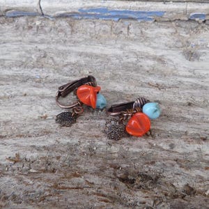May include: A pair of clip-on earrings with copper-colored metalwork. Each earring features an orange, teardrop-shaped bead, a small blue bead, and a decorative, dark-colored charm. The earrings are set against a weathered, light-colored wooden surface.