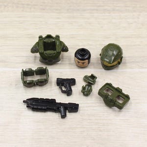 May include: A collection of olive green and black toy military accessories, including a helmet, body armor, and weapons. The set includes a rifle, a pistol, and various other components, all made of plastic.