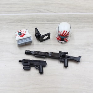 May include: A white plastic torso piece with a red handprint graphic, a black L-shaped bracket, a white helmet with a red handprint and blue visor, a long black plastic rifle, and a smaller black plastic blaster pistol toy accessory set.