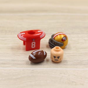 May include: A collection of miniature football-themed toys. Includes a red jersey with the number 8, a brown football, a gold helmet with a team logo, and a tan-colored minifigure head with a stern expression.