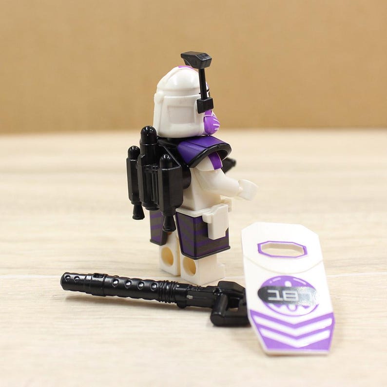 May include: A white and purple action figure with a black jetpack, weapon, and shield. The figure has a white helmet with purple accents and a black weapon. The shield is white with purple stripes and text.