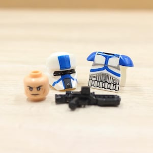 May include: A collection of toy building block pieces, including a white and blue helmet, a torso with blue accents, a black blaster, and a tan head with black eyes. The pieces are designed to be assembled.
