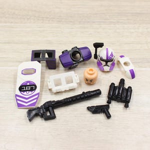 May include: A collection of toy accessories, including a purple and white helmet, a black rifle, a shield with the number "187", and other small parts. The items are arranged on a light-colored wooden surface.