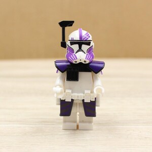 May include: A white and purple action figure of a soldier with a helmet, shoulder armor, and leg armor. The figure has a black visor and a black accessory on top of the helmet. The figure is standing on a wooden surface.