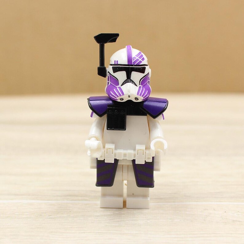 May include: A white and purple action figure of a soldier with a helmet, shoulder armor, and leg armor. The figure has a black visor and a black accessory on top of the helmet. The figure is standing on a wooden surface.