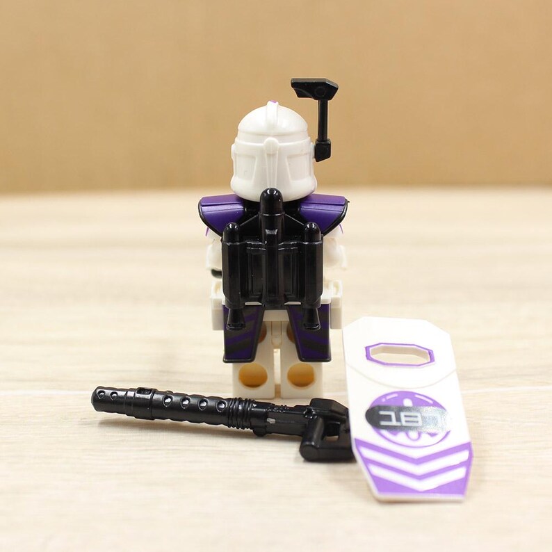 May include: A white and purple action figure with a black jetpack, helmet, and weapon. The figure has a shield with purple chevron stripes and text. The toy is a collectible item.