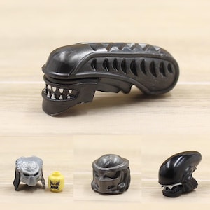 May include: Black Alien and Predator action figure heads. The image shows a black Alien head with a detailed, textured design and sharp teeth. Also included are Predator heads in gray and black, and a yellow Lego figure.