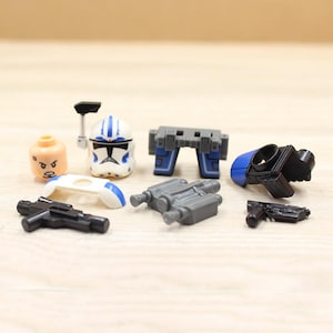 May include: A collection of miniature toy accessories, including a white and blue helmet, a tan head, gray binoculars, and black weapons. The accessories are designed for a miniature toy figure.