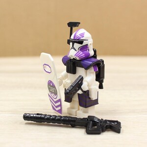 May include: A white and purple action figure of a soldier with a helmet, holding a black gun and a white shield with purple accents. A black rifle is in front of the figure. The figure has a black backpack.
