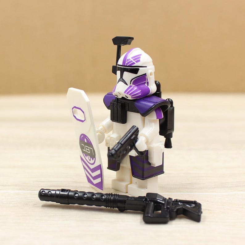May include: A white and purple action figure of a soldier with a helmet, holding a black gun and a white shield with purple accents. A black rifle is in front of the figure. The figure has a black backpack.